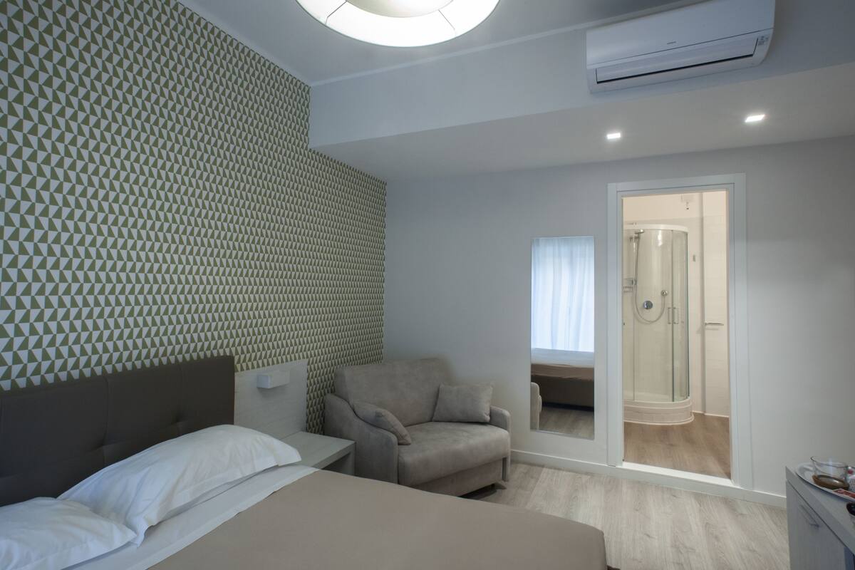 Une chambre d’hôtel moderne équipée d’un lit, d’un canapé, d’une chaise, d’une petite table et d’un espace douche visible à travers une porte en verre.