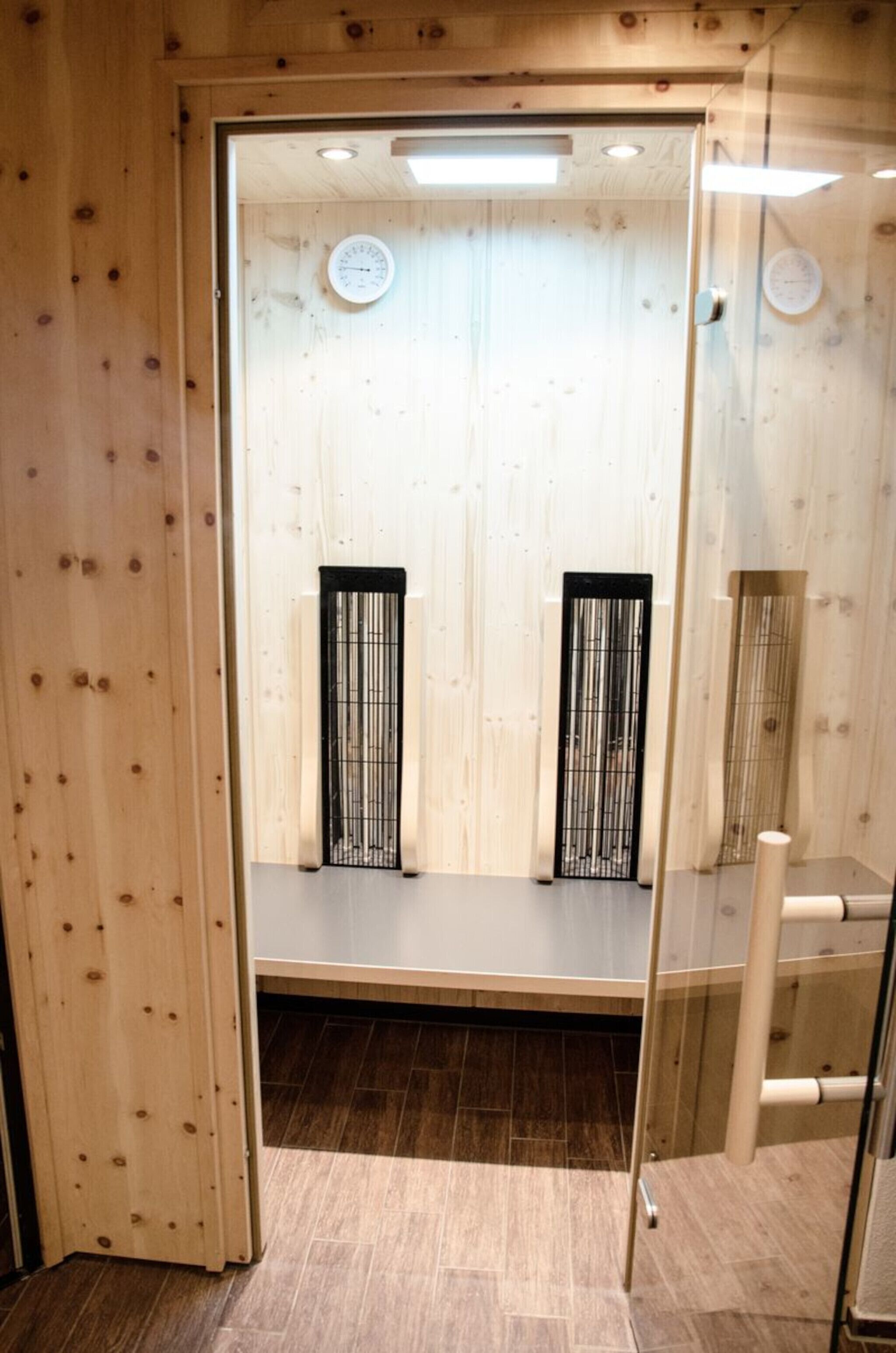 Una sala de sauna con paredes de madera, dos bancos de metal negro y una puerta de vidrio.