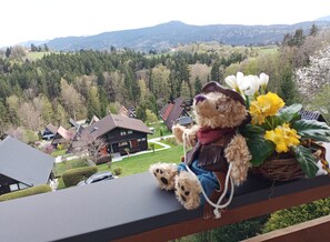 Un ours en peluche est posé sur un balcon qui donne sur un village avec des maisons traditionnelles, le tout sur fond de montagnes.