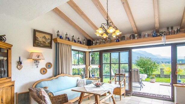 A cozy room with a blue sofa, wicker chairs, a wooden table, and a large window offering a view of the outdoors. - Ferienwohnung 1, 65 qm, f. 1-2 Personen (Grassau)