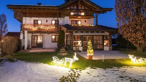 A two-story chalet with a balcony, a Christmas tree, and snow-covered ground. - Ferienwohnung 1, 65 qm, f. 1-2 Personen (Grassau)