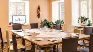 A conference room with a large table, chairs, a TV screen displaying 'Fuchs Hotels', and large windows with plants.
