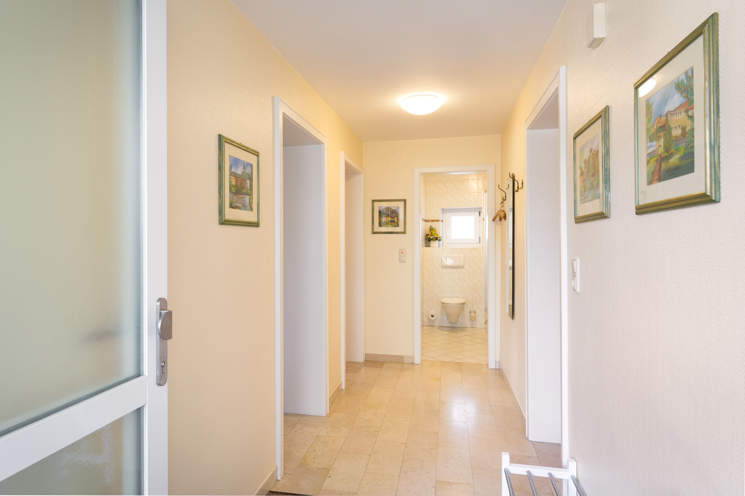 Un pasillo con puertas que dan a habitaciones, cuadros en las paredes y una lámpara en el techo.