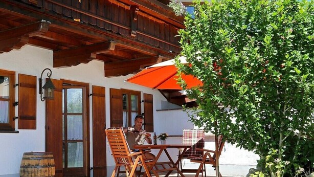 An outdoor seating area with wooden furniture and a table, a large tree, and a building with wooden beams and windows. - holiday flat (Truchtlaching)