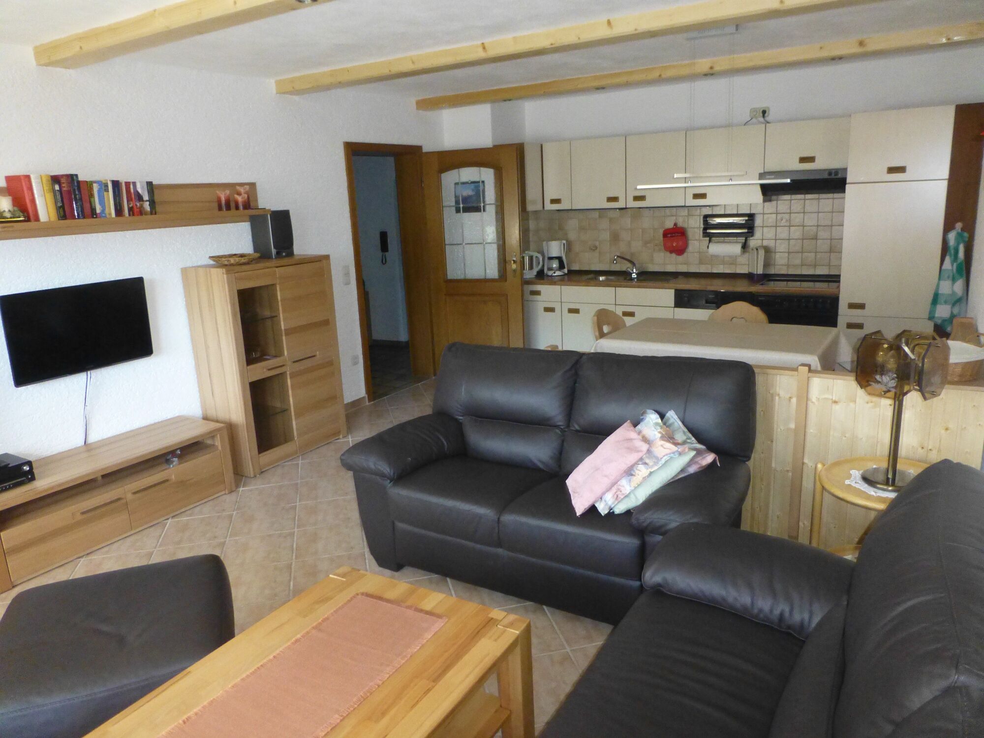 Una sala de estar con un sofá de cuero negro, una mesa de centro de madera y un televisor montado en la pared. Se ve una cocina al fondo, con armarios blancos y un salpicadero de azulejos.