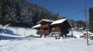 Una cabaña de madera con balcón, rodeada de árboles cubiertos de nieve y una pista de esquí.