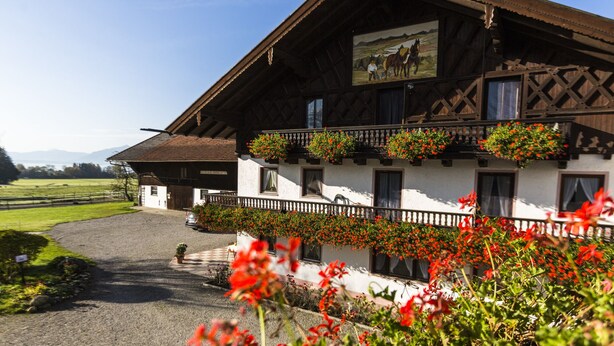 Un bâtiment de style alpin traditionnel, doté d’un balcon orné de fleurs rouges, d’une façade en bois et offrant une vue panoramique sur la campagne environnante.