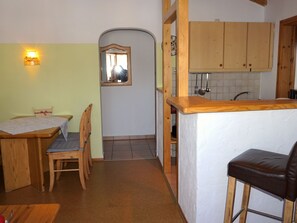A compact kitchen with wooden cabinets, a small dining area with a table and chairs, and a bar stool. - 3. Fewo Hochplatte, 38 qm, 1-4 Pers., 1 Schlafzimmer, 1 Wohnschlafraum, Balkon, 1 OG (Grassau)