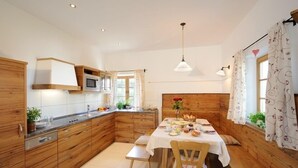 A modern kitchen with wooden cabinets, a dining table set for four, and a large window with curtains. - Ferienwohnung "Chiemsee", 110 qm Erdgeschoss, 3 separate Schlafzimmer (Höslwang)