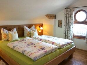 Una cama doble con ropa de cama floral, cabecera de madera, lámpara de noche y ventana con cortinas.