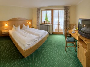 A hotel room with a large bed, wooden headboard, two bedside tables with lamps, a desk with a chair, a television, and a window with curtains.