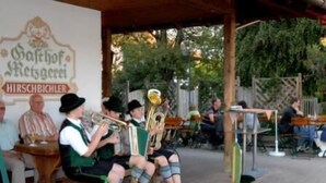 Um grupo de músicos tocando ao ar livre, sentados em bancos de madeira sob uma área coberta com um letreiro acima.