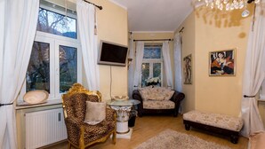 Een gezellige woonkamer met een fauteuil met luipaardprint, een vintage bank en een klein bankje. Een televisie is aan de muur bevestigd, en er zijn twee ramen met gordijnen.