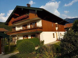 A two-story chalet with a balcony, surrounded by lush greenery and mountains in the background.