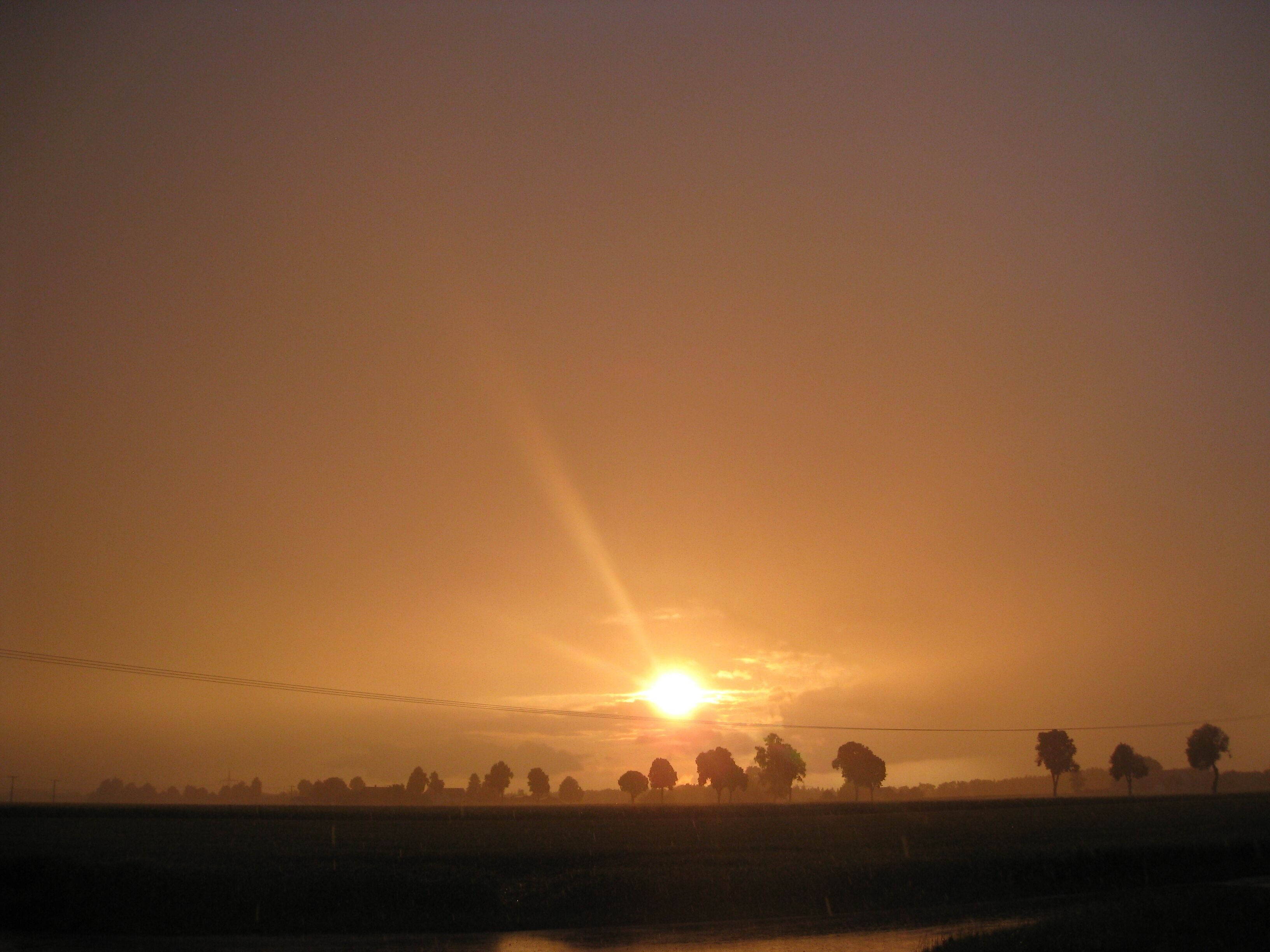 Ein Sonnenuntergang über einer ländlichen Landschaft, Felder und Bäume zeichnen sich vor dem Himmel ab.