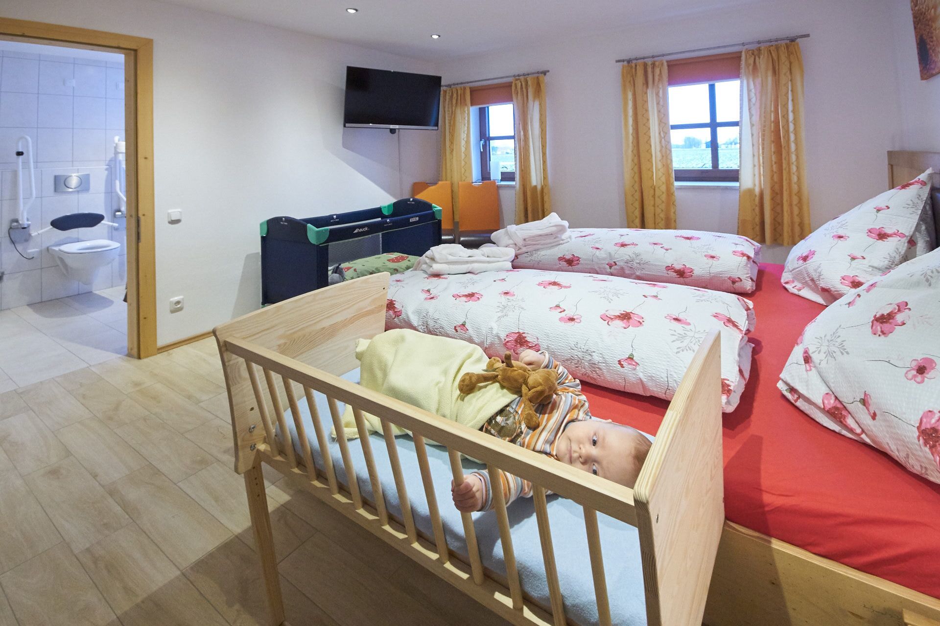 Een kamer met een babybedje, een bed met bloemenbeddengoed, een televisie en een raam met gordijnen.