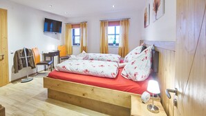 A bedroom with a wooden bed, a desk, a chair, a TV, and two windows with curtains.