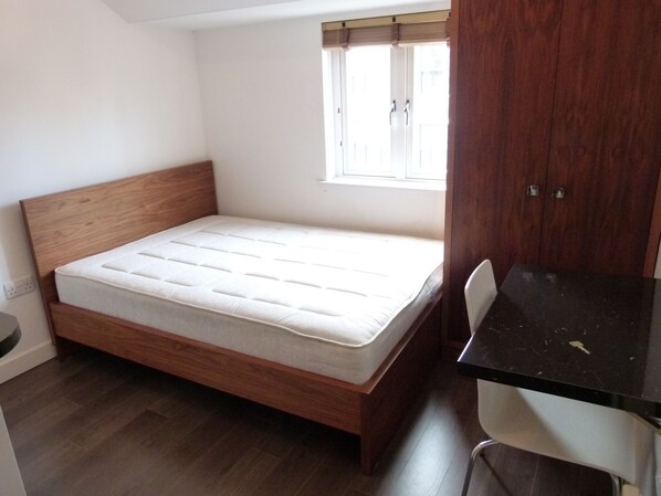 A small, single-bed room with a wooden headboard, a window with blinds, a desk with a chair, and a wardrobe.