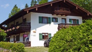A traditional chalet-style building with a balcony, a car parked in front, and lush greenery surrounding it.