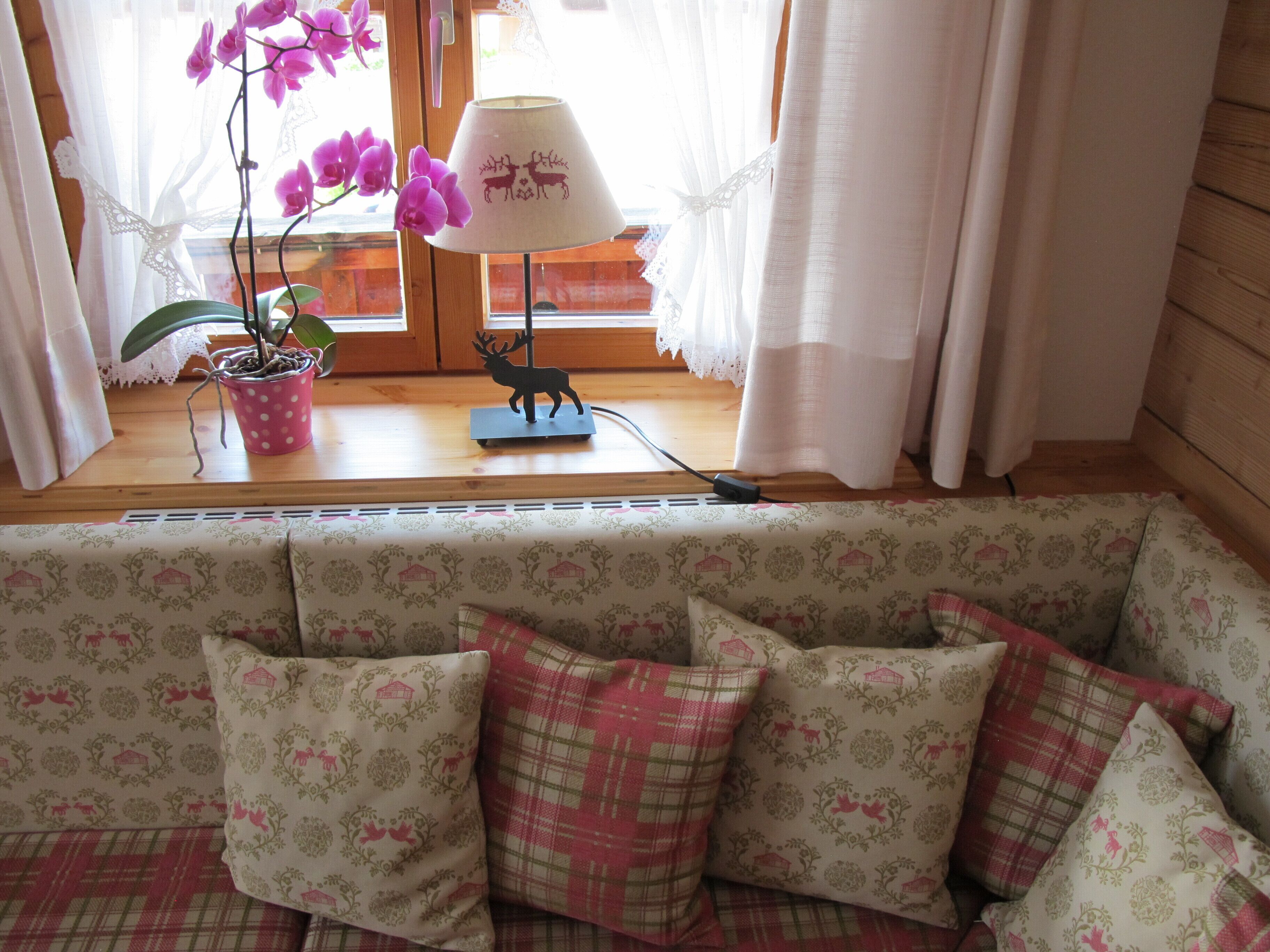 Un salon chaleureux avec un canapé à motifs, une orchidée en pot, une lampe dont l’abat-jour est orné de la silhouette d’un cerf, et une fenêtre avec des rideaux.