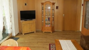 A living room with wooden paneling, a television on a cabinet, a glass-fronted cabinet, and a door. - holiday flat "Traunstein" (80 qm), balcony, kitchen, 3 bedrooms, 1 livingroom, max. 6 persons (Ruhpolding)