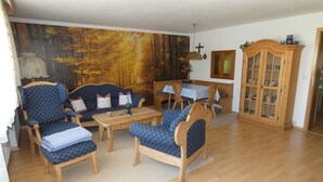 A cozy living room with wooden furniture, a large forest-themed wall mural, and a wooden cabinet with glass doors. - holiday flat "Traunstein" (80 qm), balcony, kitchen, 3 bedrooms, 1 livingroom, max. 6 persons (Ruhpolding)