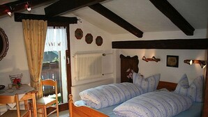 A bedroom with a wooden bed, a table with chairs, a radiator, and a window with curtains. - Zwei-Raum-Appartement (5) 45qm, Extra-Schlafzimmer mit Dusche/WC, Miniküche, Balkon (Reit im Winkl)