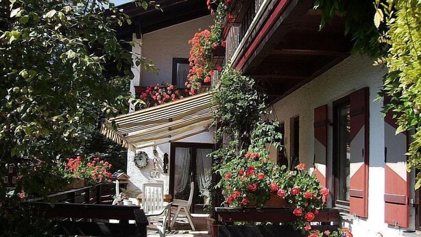 A traditional building with a balcony adorned with red flowers, a wooden pergola, and outdoor seating. - Zwei-Raum-Appartement (5) 45qm, Extra-Schlafzimmer mit Dusche/WC, Miniküche, Balkon (Reit im Winkl)
