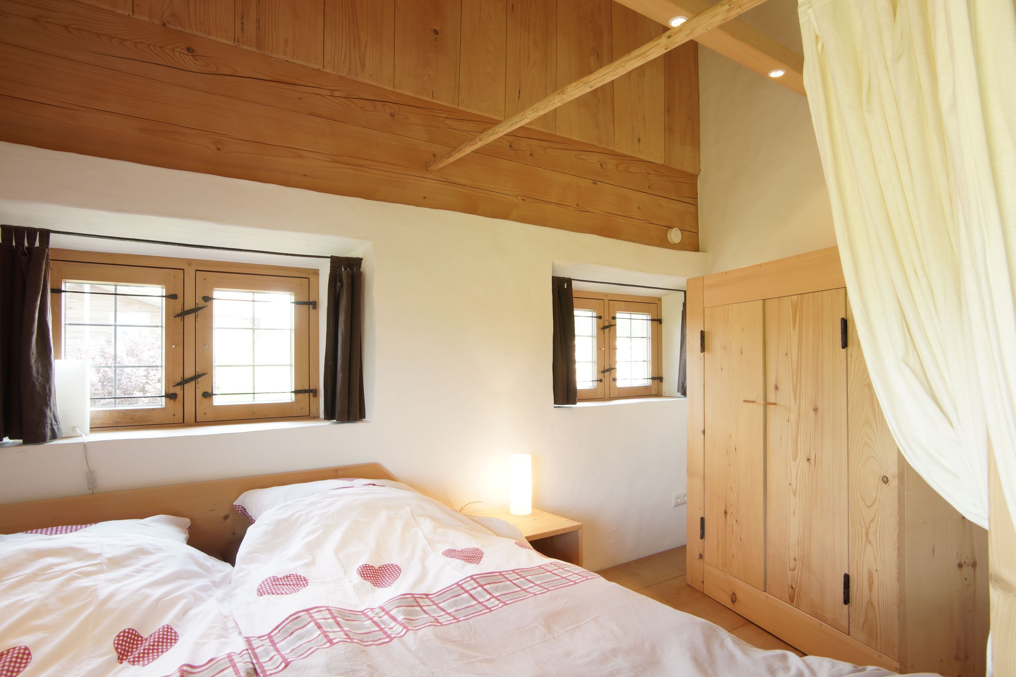 Ein Schlafzimmer mit Holzbalkendecke, zwei mit Vorhängen drapierten Fenstern, einem Bett mit herzenmusternder Bettwäsche, einem Nachttisch mit Lampe und einem Kleiderschrank.