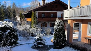 Een besneeuwde tuin met een houten huis, een verhard pad en sneeuwzware bomen.