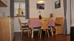 Een eetruimte met een houten tafel en stoelen, een rood-wit geruit tafelkleed, een hangende hanglamp en een raam met gordijnen.