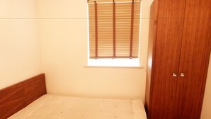 A bedroom with a wooden bed frame, a window with blinds, and a wooden wardrobe. - SS Property Hub - City of London Studio Apartment 18 (London)