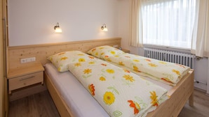 Un letto matrimoniale con biancheria da letto floreale, due lampade da comodino, un comodino in legno con un cassetto e una finestra con tende trasparenti.