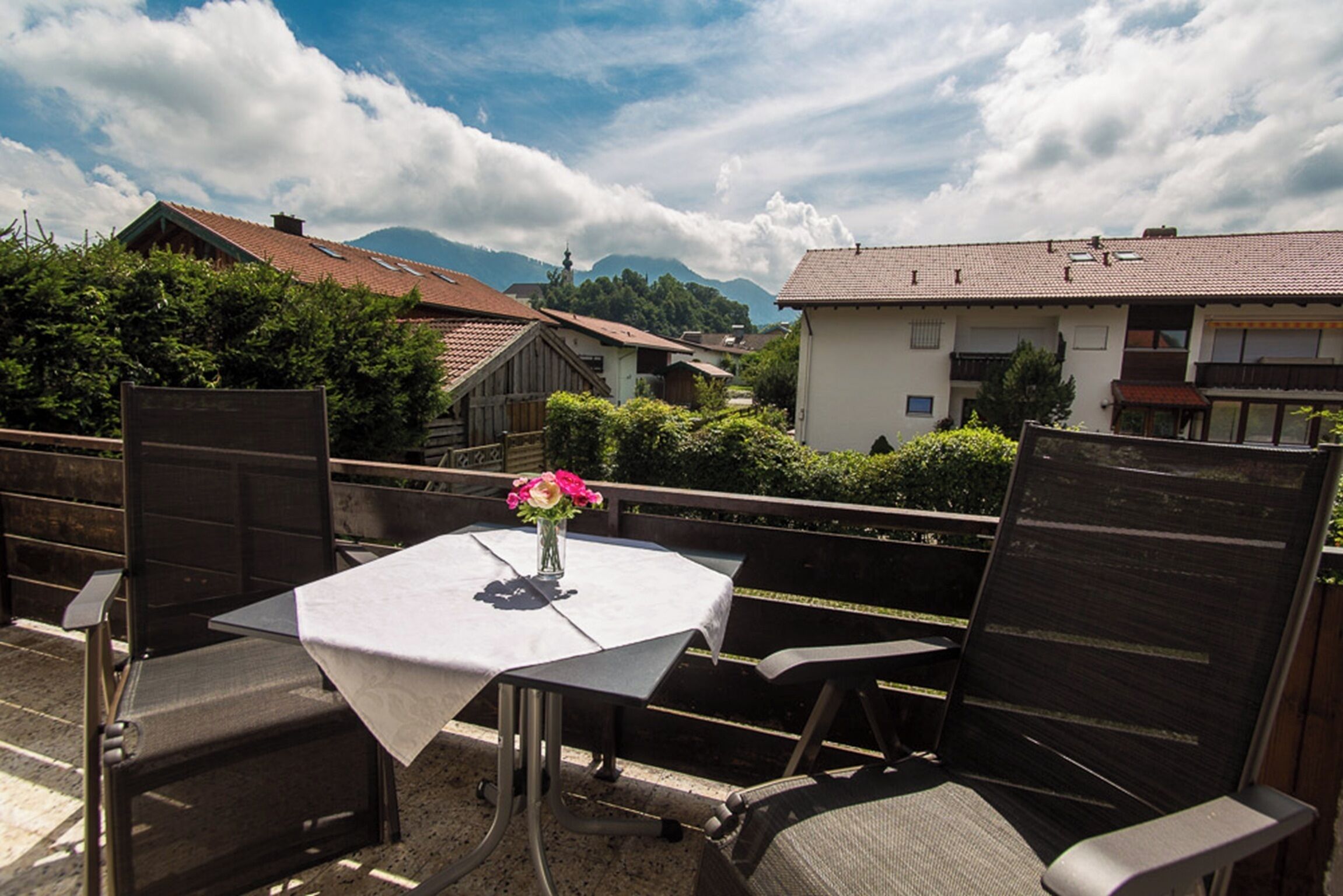 Une terrasse extérieure avec une table et des chaises, un vase rempli de fleurs et, en arrière-plan, une vue sur des maisons et des montagnes.