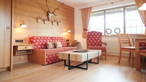A cozy room with a wooden wall, a sofa with a red and white pattern, a wooden table, and a window with curtains. - Two-room apartment 5, approx. 40sqm with spacious balcony and mountain view (Reit im Winkl)