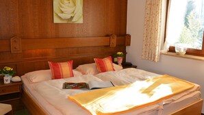 A bedroom with a wooden headboard, a bed with white bedding and red pillows, a bedside table with a vase of flowers, and a window with curtains. - Ferienwohnung Nr. 3 Untersbergblick, 1 bis 2 Personen, 45 qm, 1 Schlafzimmer, Wohnraum mit Schlafcouch, Balkon (Schönau am Königssee)