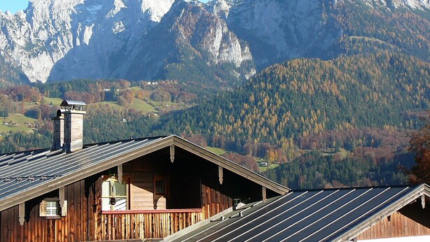 A wooden cabin with a sloped roof and a chimney, set against a backdrop of snow-capped mountains and a forested valley. - Ferienwohnung Nr. 3 Untersbergblick, 1 bis 2 Personen, 45 qm, 1 Schlafzimmer, Wohnraum mit Schlafcouch, Balkon (Schönau am Königssee)