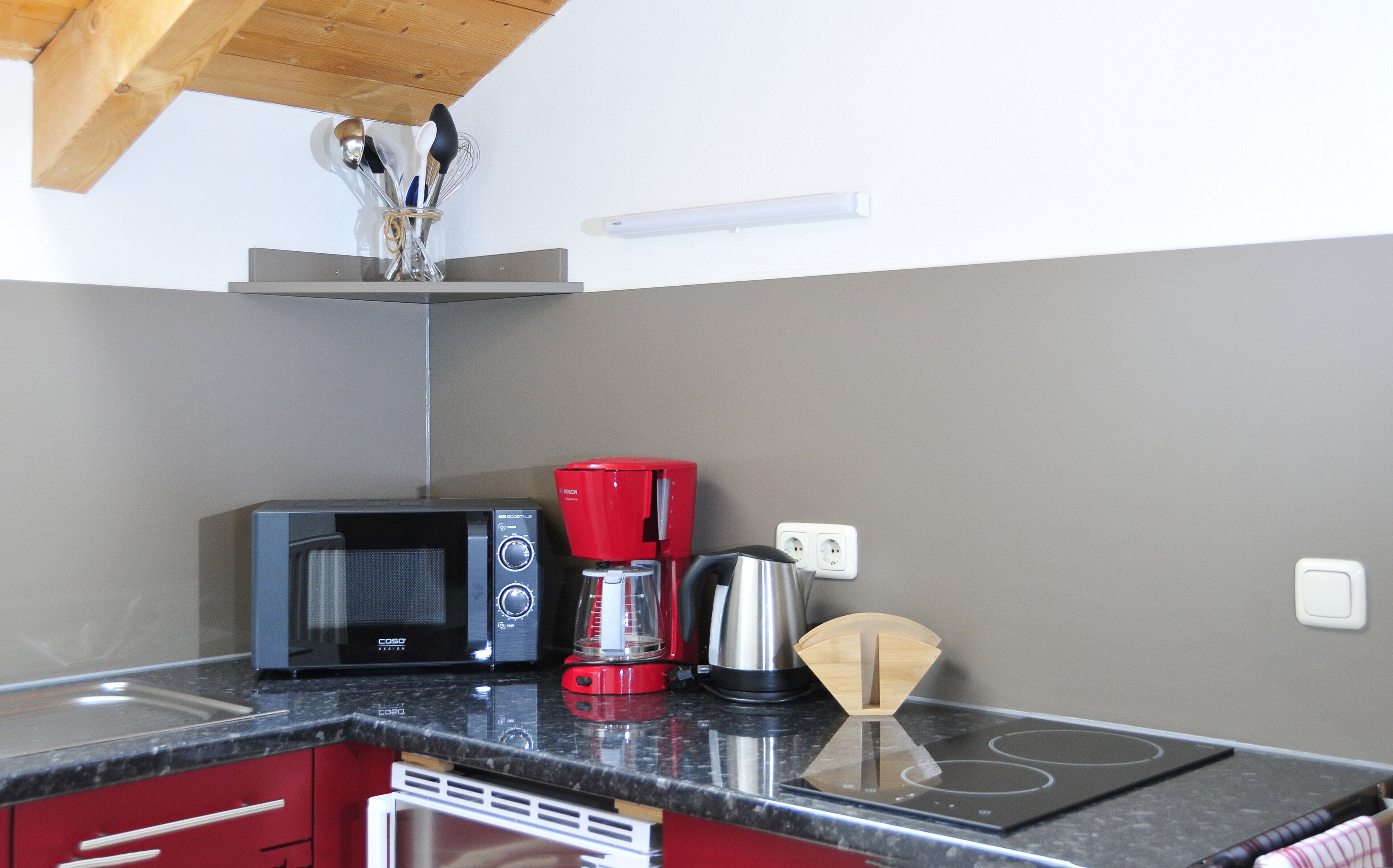 Une cuisine équipée d’un four à micro-ondes, d’une cafetière rouge, d’une bouilloire et d’une planche à découper en bois.