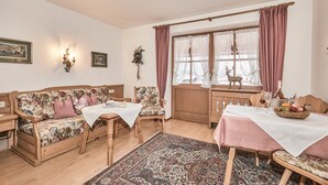 Una sala de estar acogedora con un sofá de madera, una mesa de comedor con mantel rosa y un ventanal con cortinas.