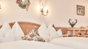 Una habitación de hotel con cabecera de madera, ropa de cama blanca, almohada con motivos florales, lámpara de pared, decoración de cabeza de ciervo y un cuadro enmarcado.