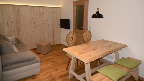 A cozy room with a wooden floor, a sofa, a wooden dining table with chairs, a small stool, and a television mounted on the wall.