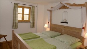 A room with two beds, a wooden headboard, a window with curtains, a bedside table with a lamp, and a small shelf with a decorative item.