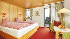 Ein Zimmer mit einem hölzernen Bett, rot-weiß karierter Bettwäsche, einem grünen Sessel, einem Tisch mit Früchten und Blick durch ein Fenster mit Vorhängen.
