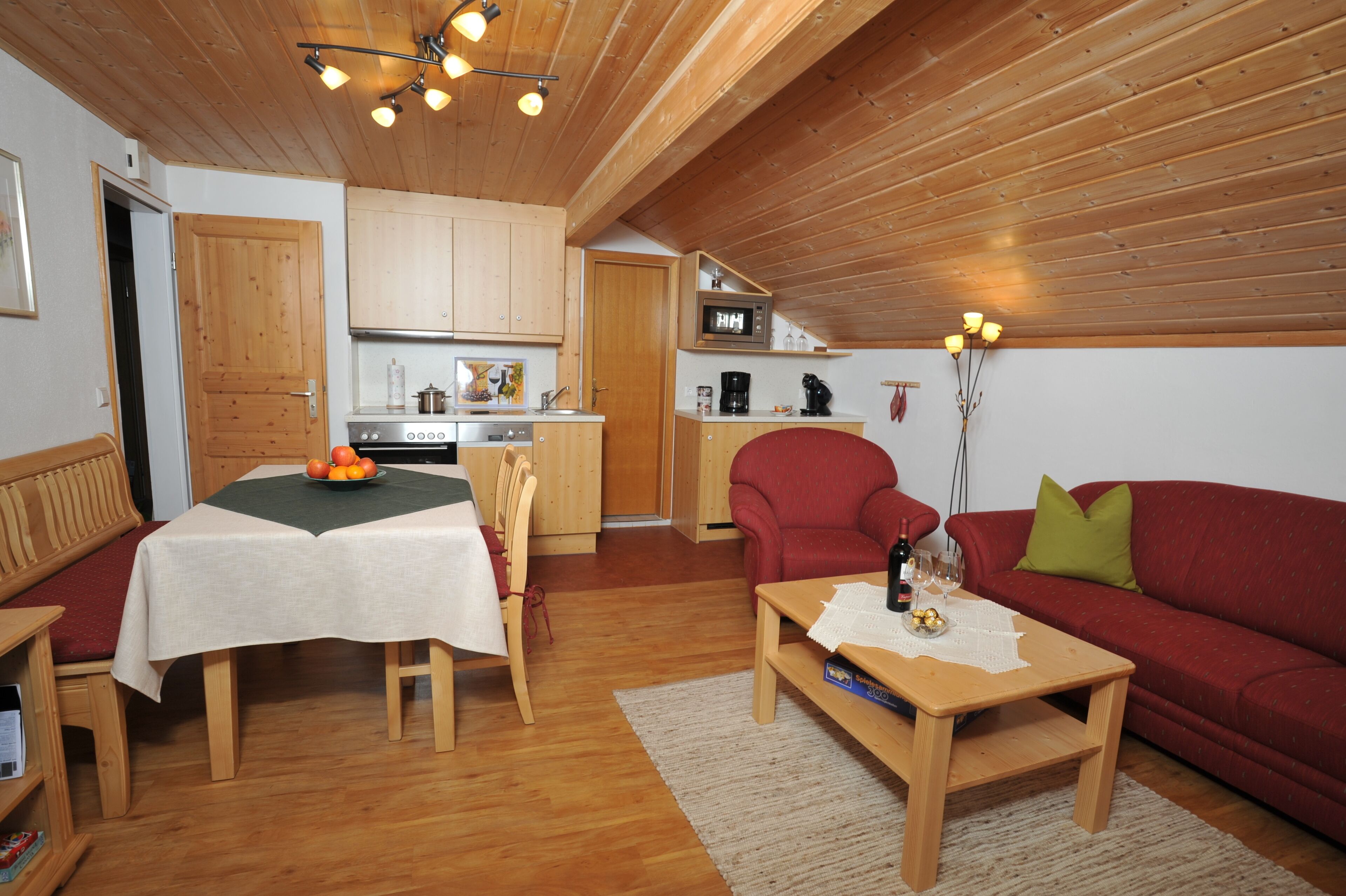A cozy living room with a wooden ceiling, a dining table with a white tablecloth, a red sofa, and a kitchen area with wooden cabinets and a microwave.