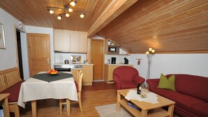 Ein gemütliches Wohnzimmer mit Holzbalkendecke, einem Esstisch mit weißem Tischlein, einem roten Sofa und einer Küchenzeile mit Holzschränken und einer Mikrowelle.