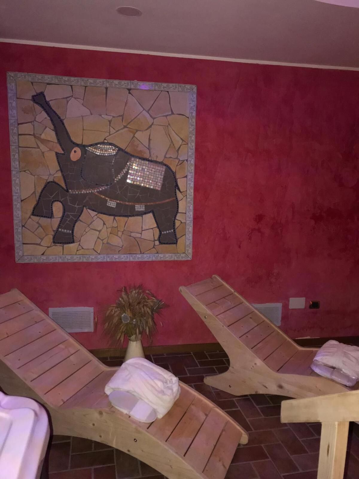 A room with a mosaic wall art of an elephant, wooden lounge chairs with towels, and a potted plant.