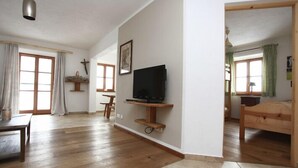 A spacious living room with a wooden floor, a television on a wooden stand, a dining table, and a bed in the adjacent room. - Vacation apartment Sophie, 70 sqm, first floor, 2 bedrooms (Schechen)