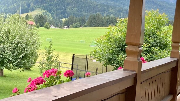 Uitzicht vanaf een balkon met houten spijlen, op een groen veld en een bergketen.