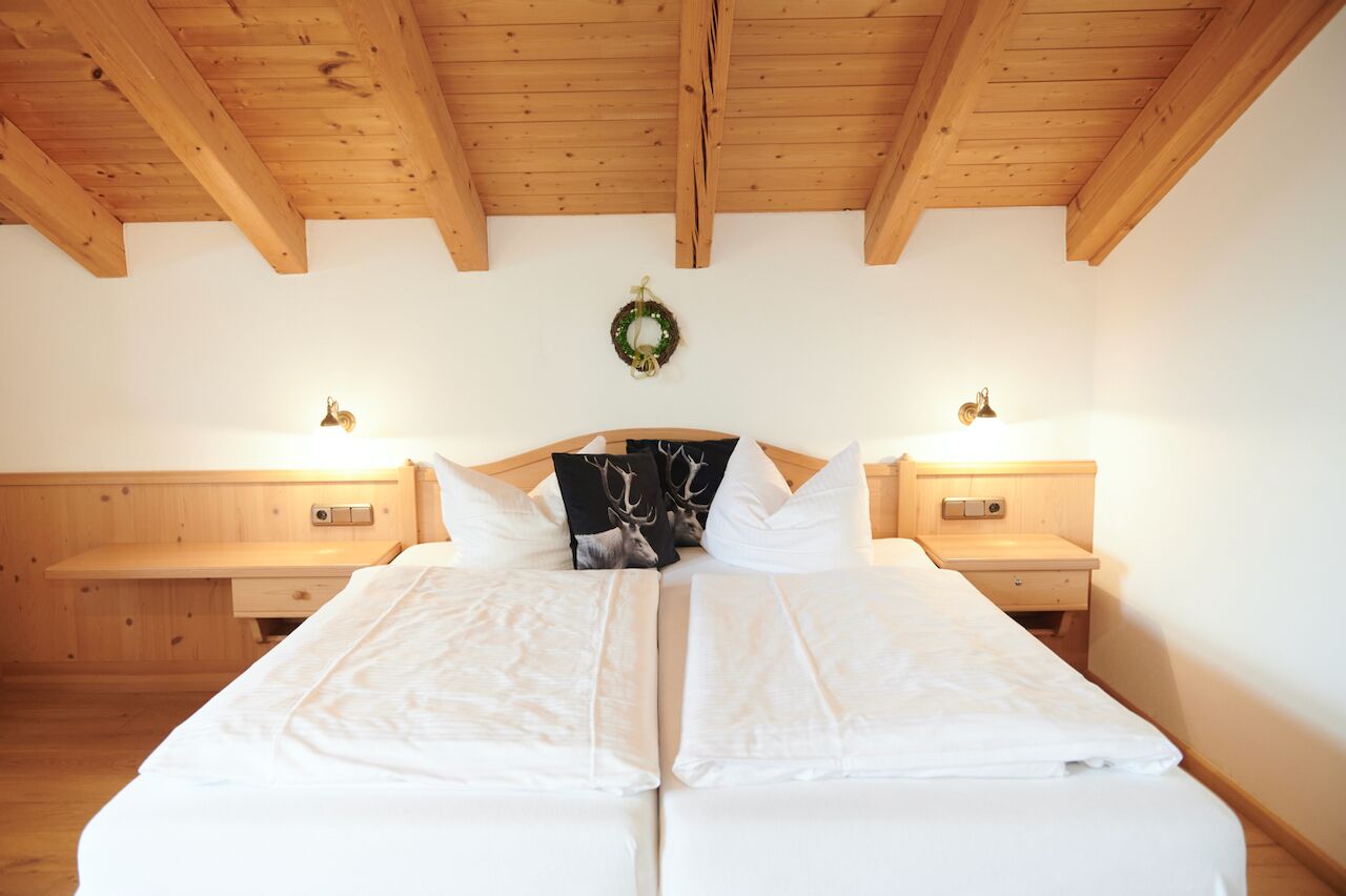 A double bed with white linens, two bedside tables with lamps, a wooden ceiling, and a wall-mounted wreath.