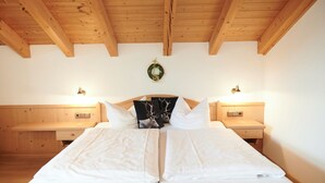 Uma cama de casal com roupa de cama branca, duas mesas de cabeceira com abajures, teto de madeira e um ramo decorativo fixado na parede.